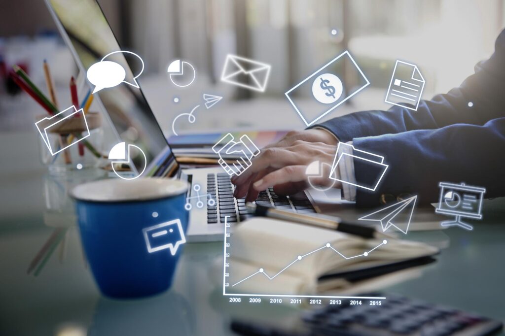This image shows a person working on a laptop at a desk, suggesting a business or office environment. Around the person’s hands, there are floating digital icons like charts, email symbols, a dollar sign, a document, a folder, and a paper airplane.