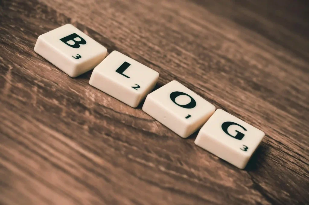 This image shows four Scrabble-like tiles arranged on a wooden surface, spelling out the word "BLOG". Each tile has a letter (B, L, O, G) and a small number below it, just like in the game Scrabble, indicating the letter's point value (B - 3, L - 2, O - 1, G - 3). The background is a wooden texture, giving the image a warm, rustic feel